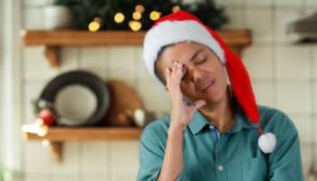 Woman wearing Santa hat standing in decorated kitchen holding head in hands, looking tired and stressed