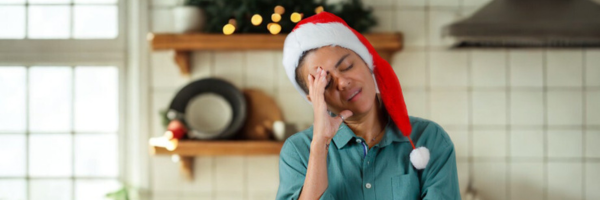 Woman wearing Santa hat standing in decorated kitchen holding head in hands, looking tired and stressed