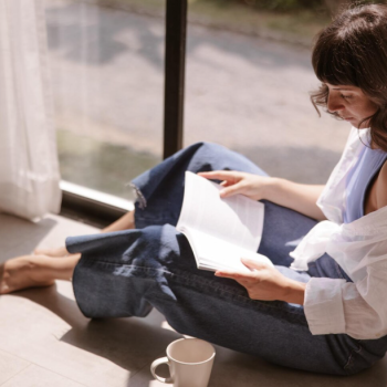 Person sitting on the floor by a sunny window reading a book with a coffee mug nearby.