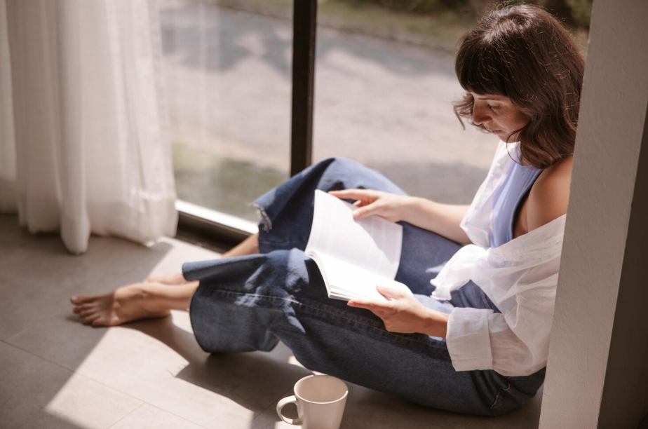 Person sitting on the floor by a sunny window reading a book with a coffee mug nearby.